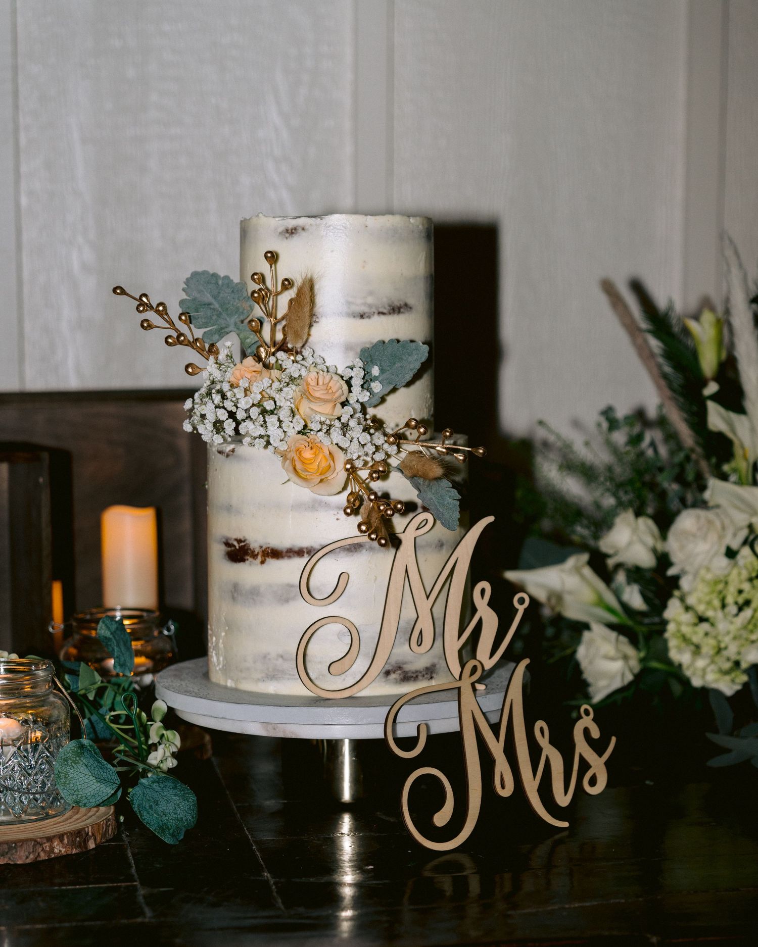Rustic wedding cake with floral accents and a “Mr & Mrs” topper, displayed on a table with soft lighting.
