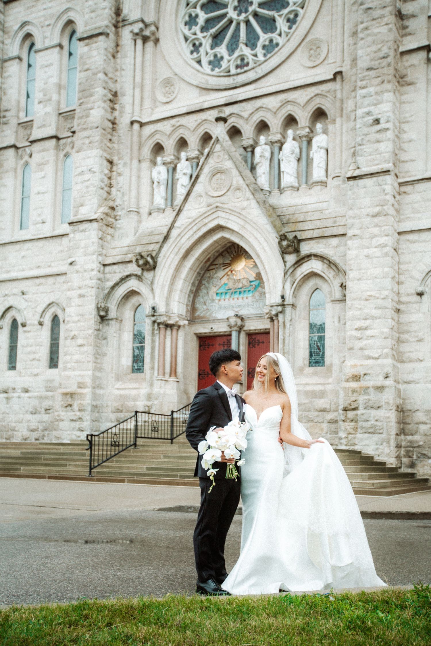Wedding couple poses in front of gothic stone church with ornate architecture.