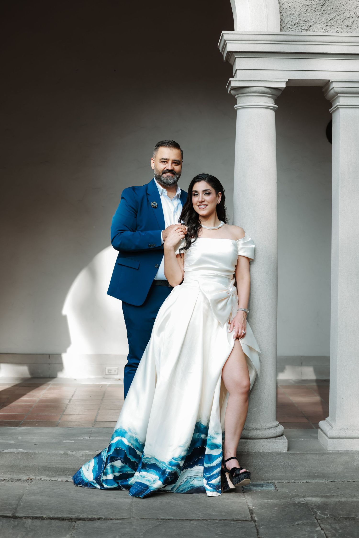 A couple poses against white classical columns in formal wedding attire.