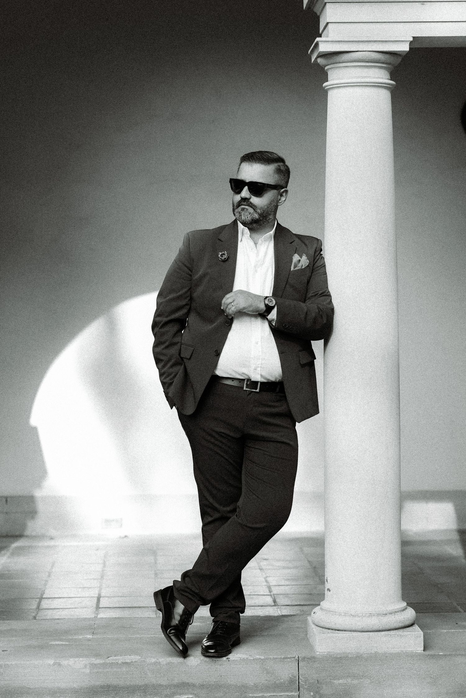 Monochrome portrait of suited man with sunglasses leaning against white column.