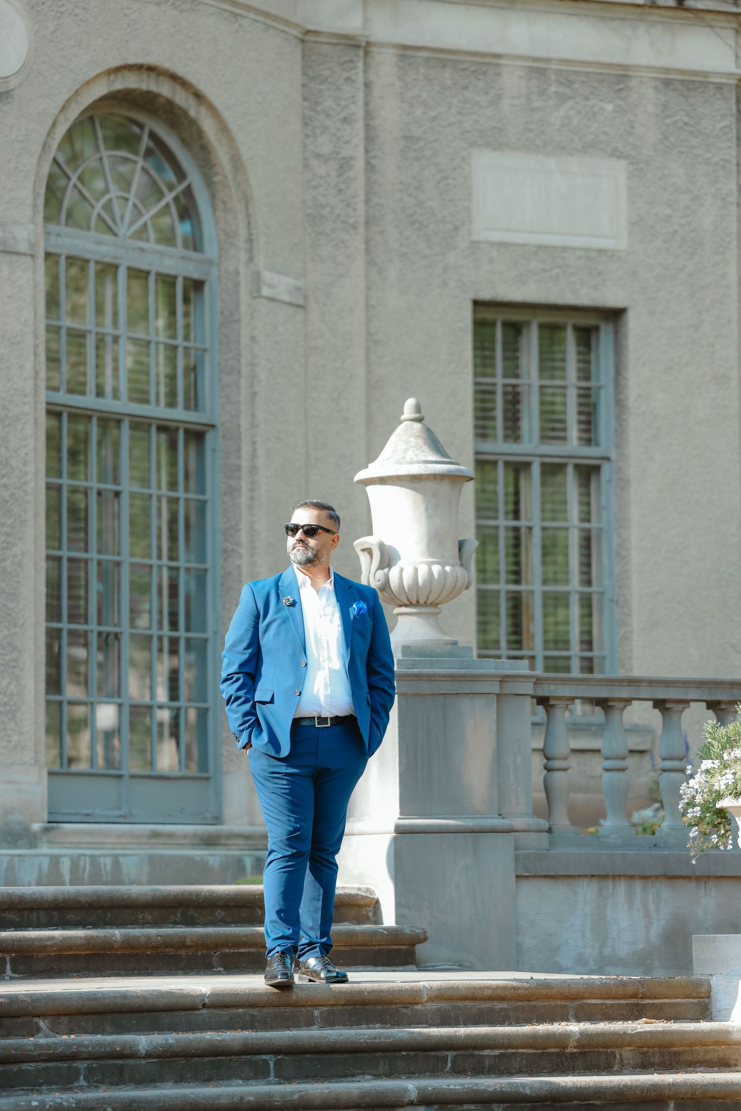 Person in blue suit stands confidently on stone steps near decorative urn and arched windows.