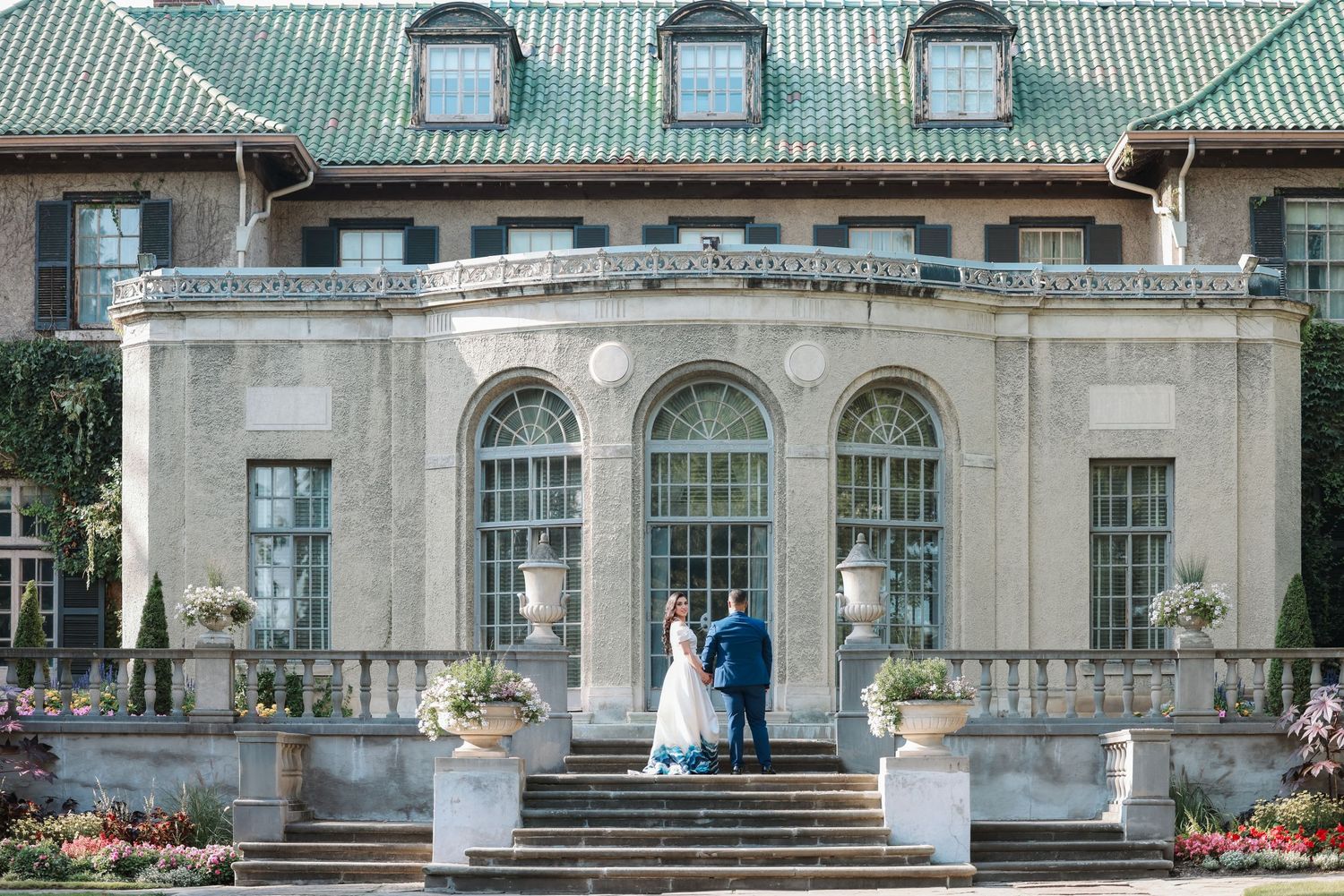 A wedding couple poses on the steps of a grand historic building with green roof and arched windows.