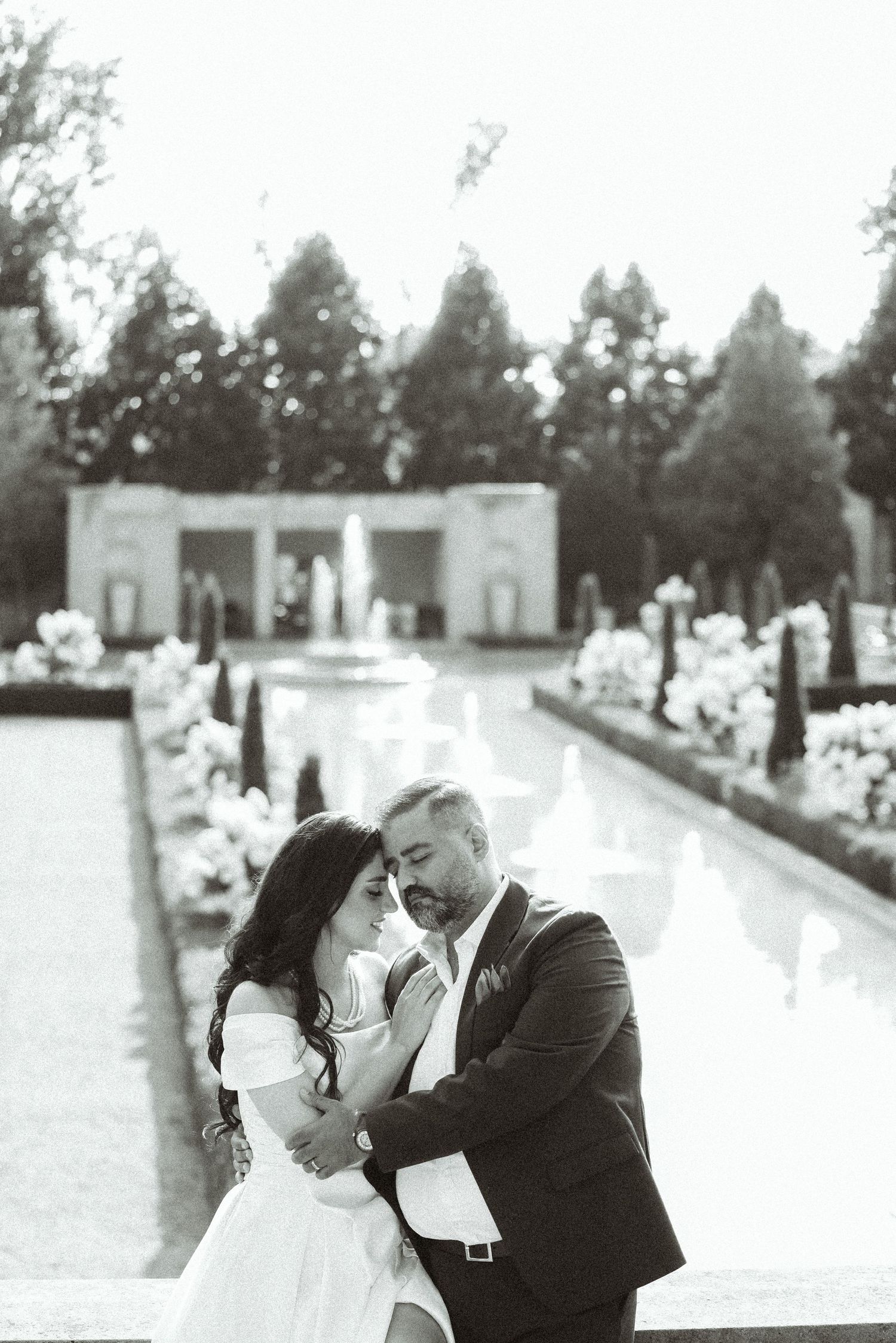 Black and white portrait of newlyweds in an elegant garden setting with water features and columns.