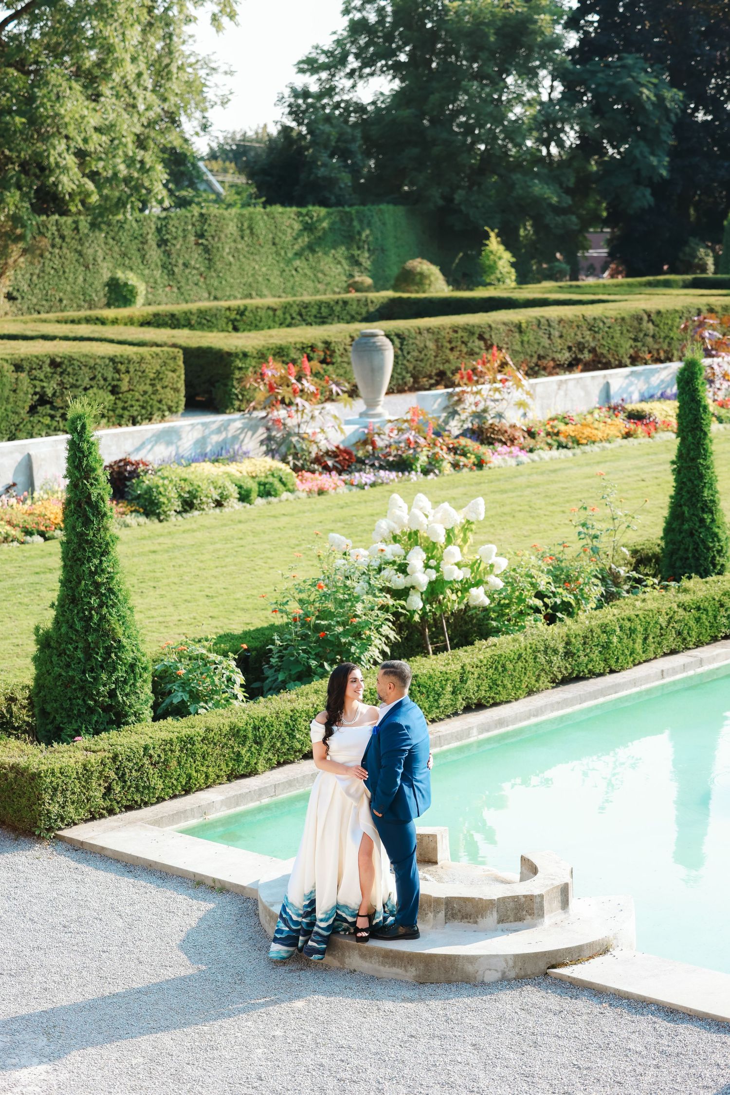 Formal garden wedding photo with couple by pool surrounded by trimmed hedges and colorful flowers.