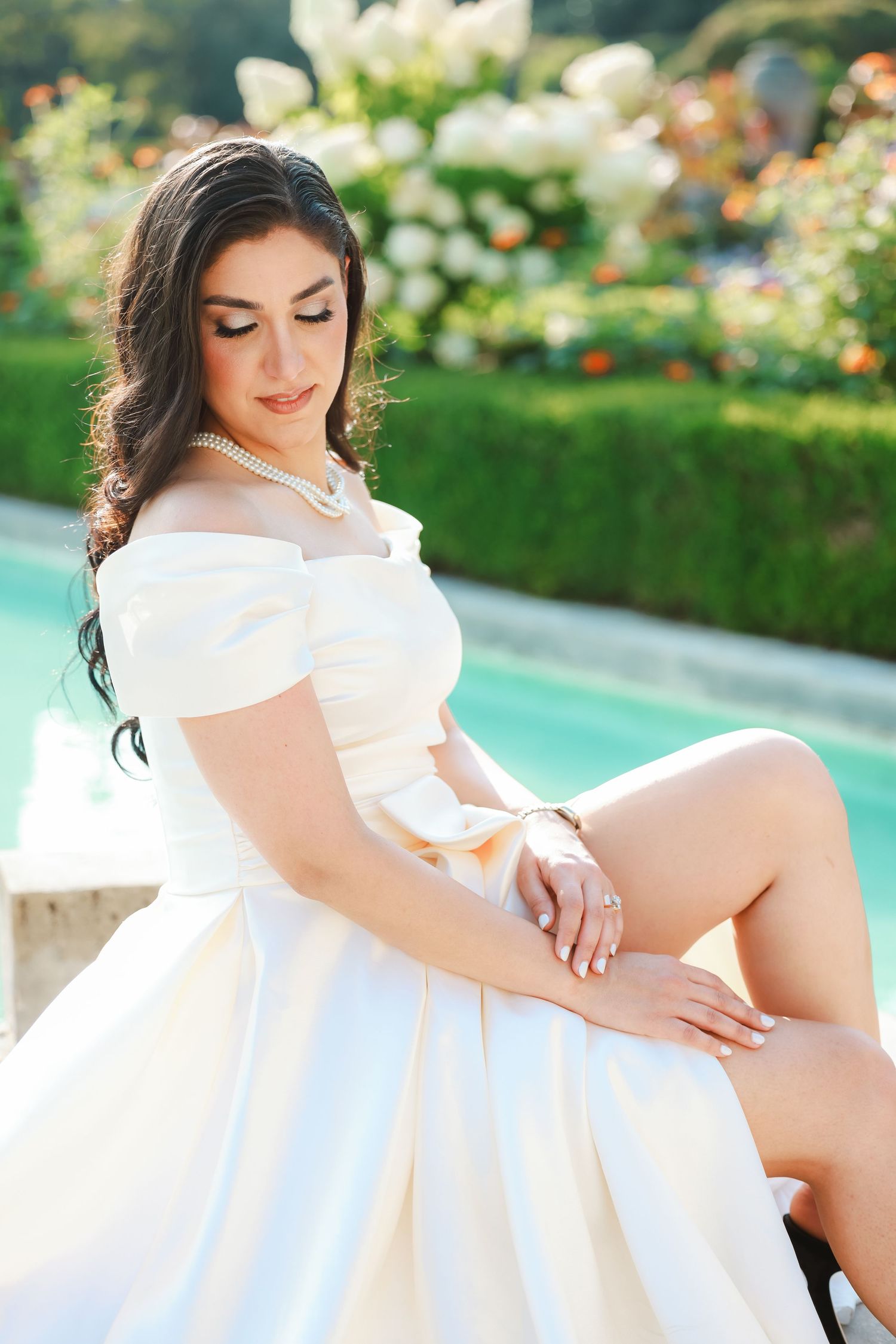 Bride in off-shoulder white dress poses by pool in garden setting.