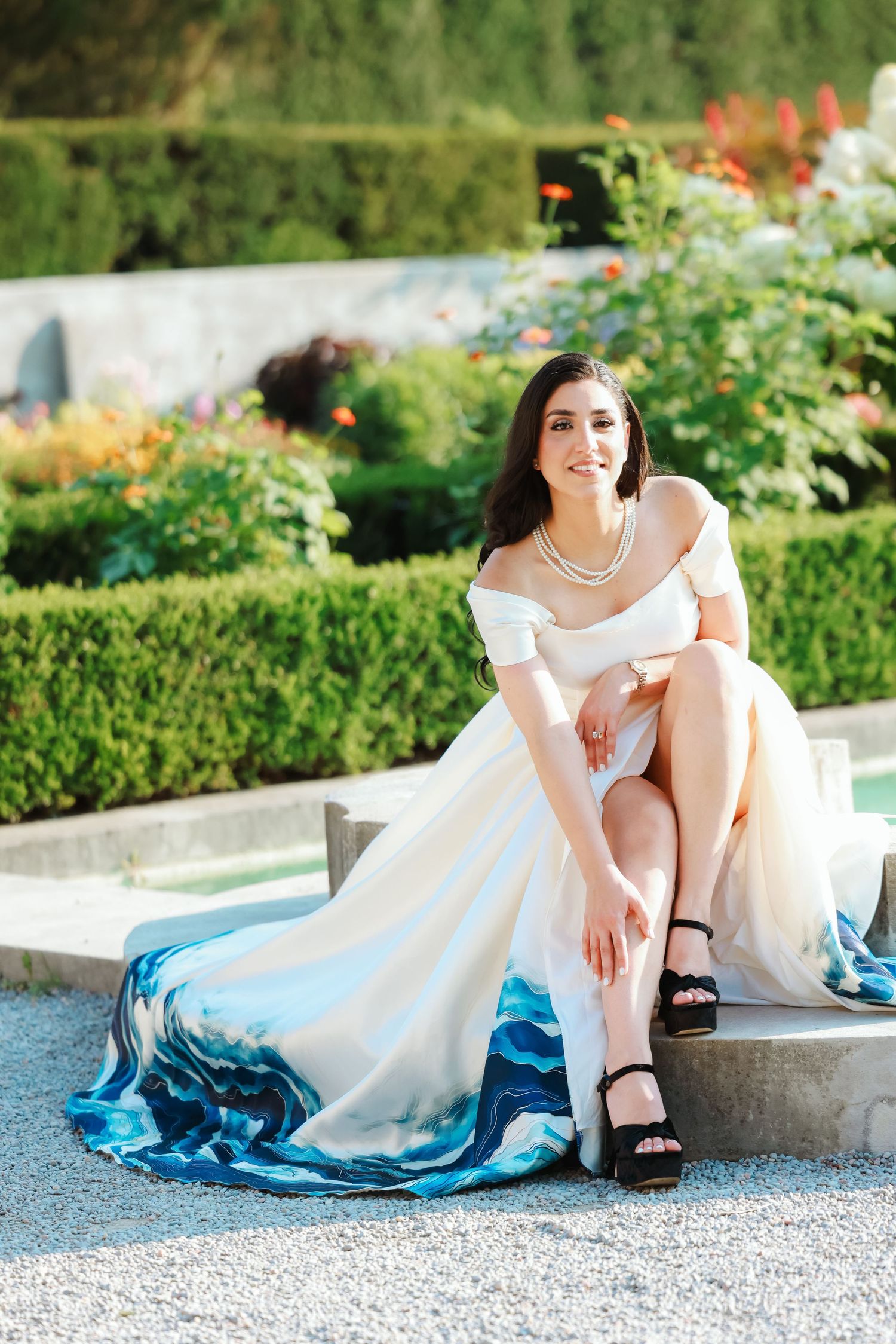A white off-shoulder dress with blue ombre hem flows gracefully as someone poses on garden steps.