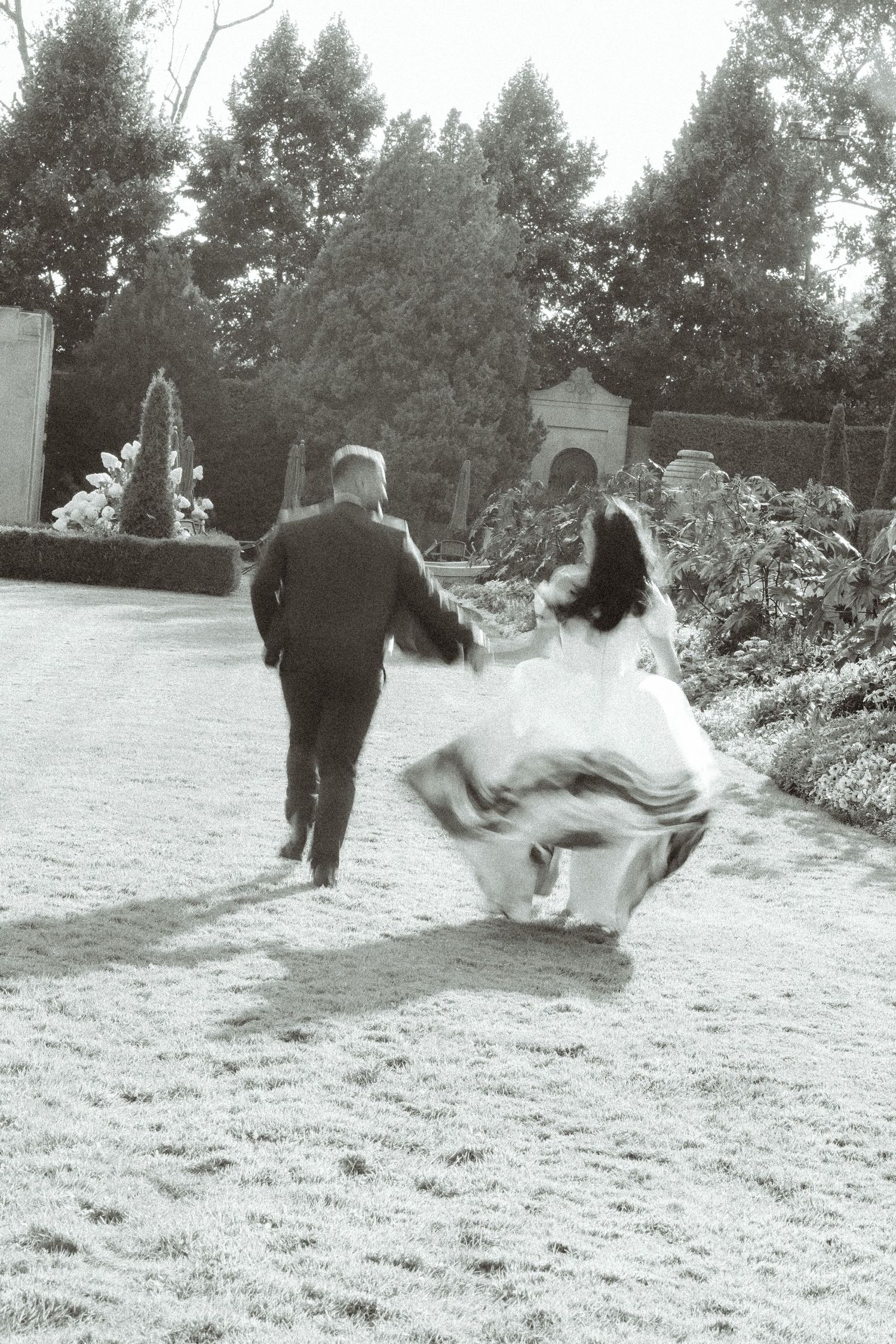A black and white photo captures movement as two people run through a garden pathway.