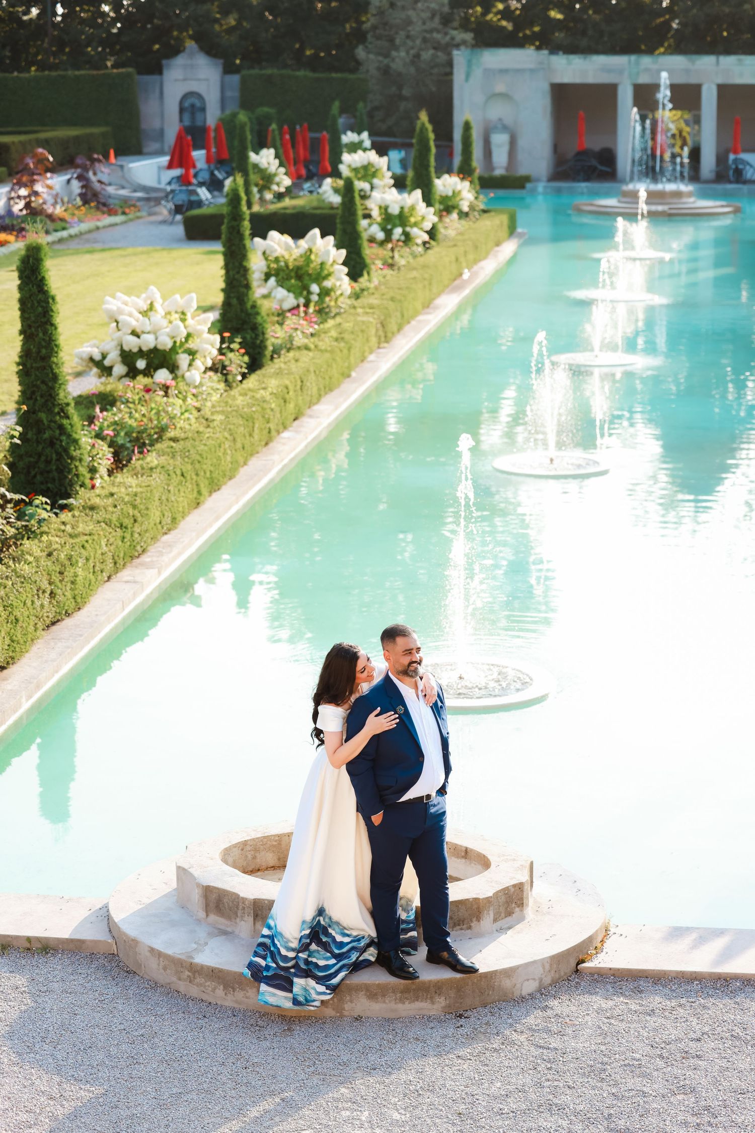 A couple shares a romantic moment by an elegant pool with fountains in a formal garden setting.