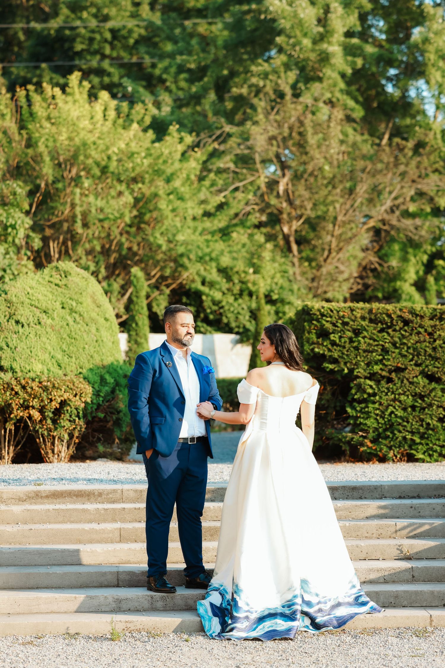 Couple in wedding attire stands on stone steps surrounded by manicured garden hedges.
