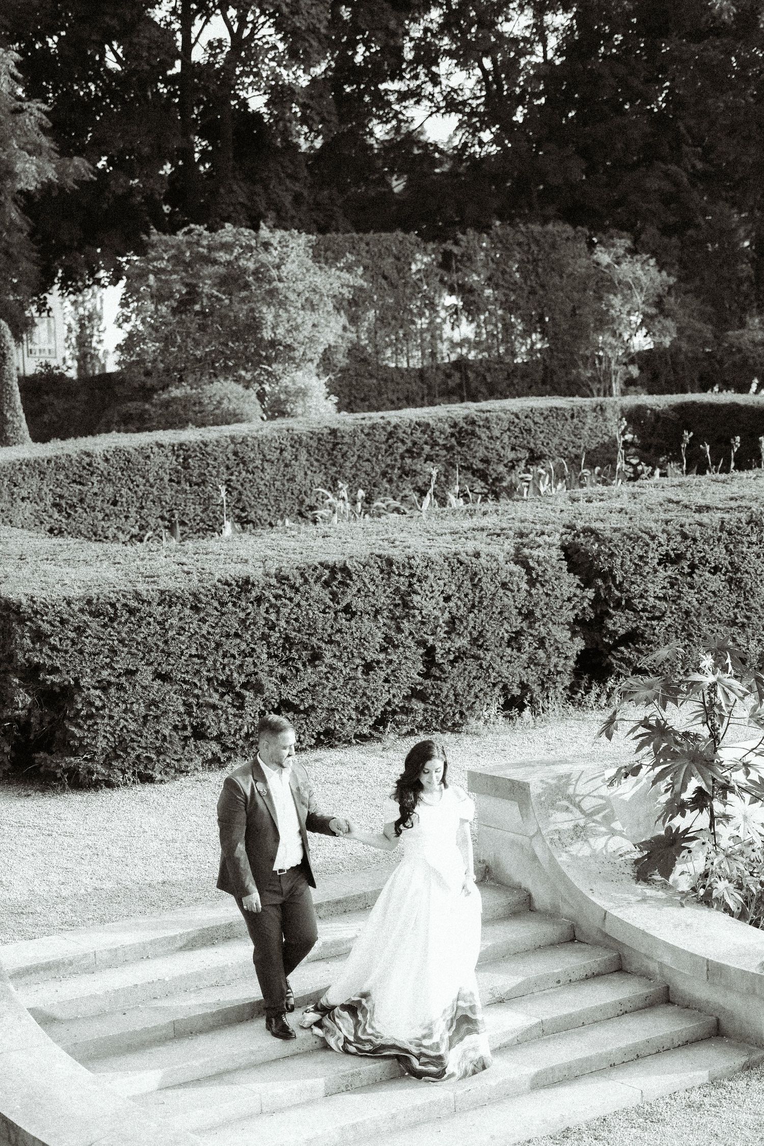 A couple descends ornate garden steps surrounded by manicured hedges in a black and white wedding photo.