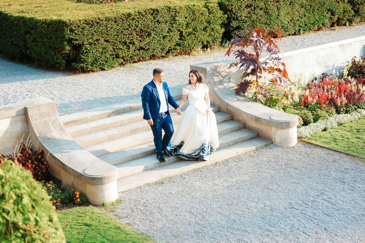 Newlyweds walking down ornate garden steps with red flowers and hedges.