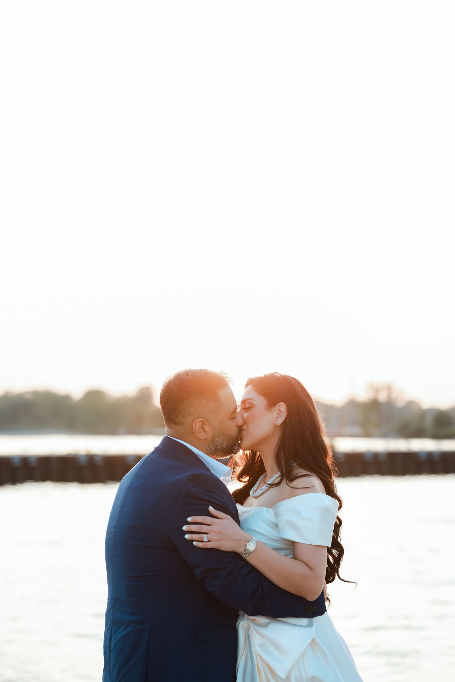 A romantic couple embraces and kisses on a waterfront dock during sunset in wedding attire.