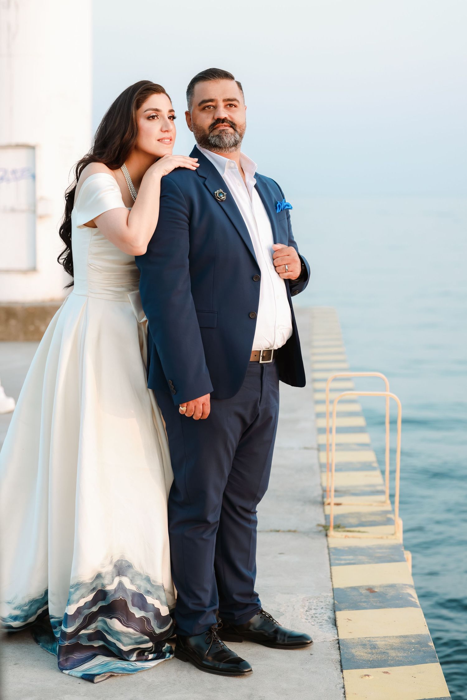 Wedding couple standing on waterfront pier with bride's dress featuring unique wave-pattern detail.