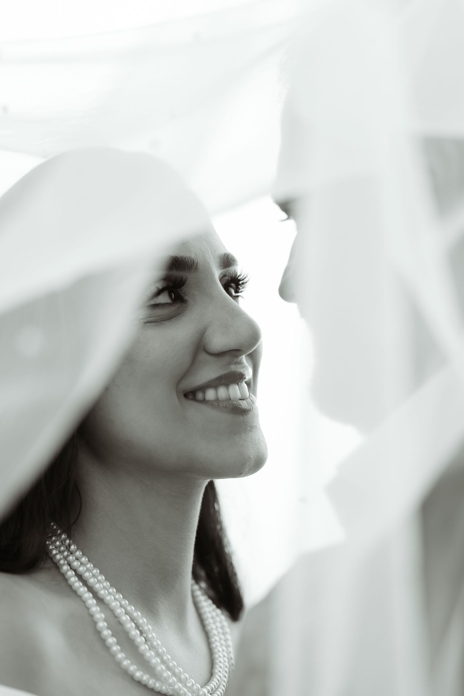 Black and white portrait of person smiling under veil wearing pearl necklace.