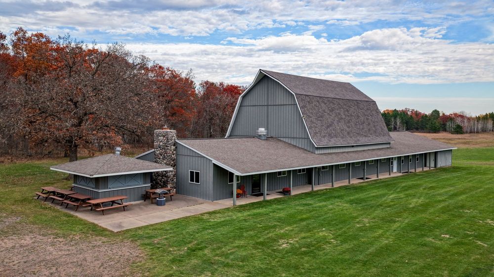 Gray barn-style lodge with a gambrel roof, stone chimney, covered porch and picnic tables beside a green lawn and autumn trees.