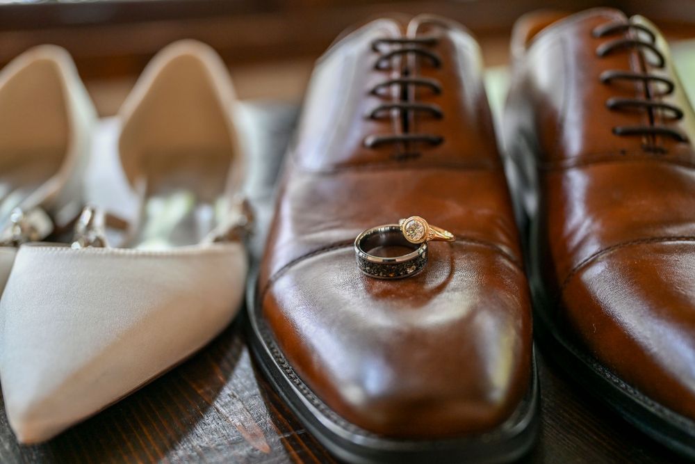 Wedding rings resting on brown leather Oxford dress shoes beside white bridal heels on a wooden surface.