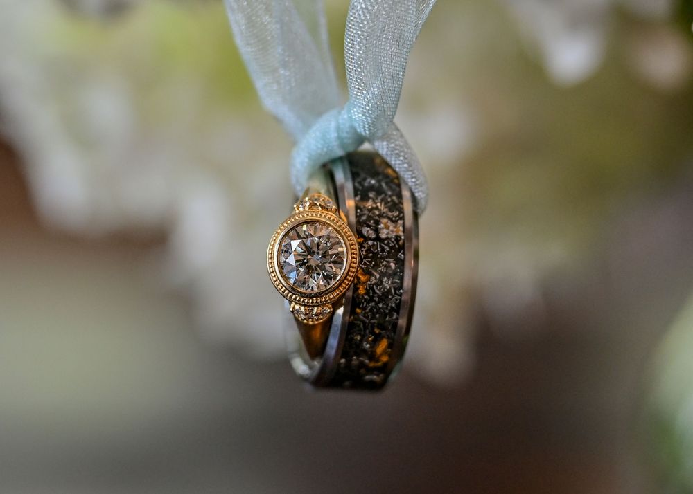 Elegant wedding rings tied with a sheer blue ribbon, featuring a diamond engagement ring and dark patterned band.