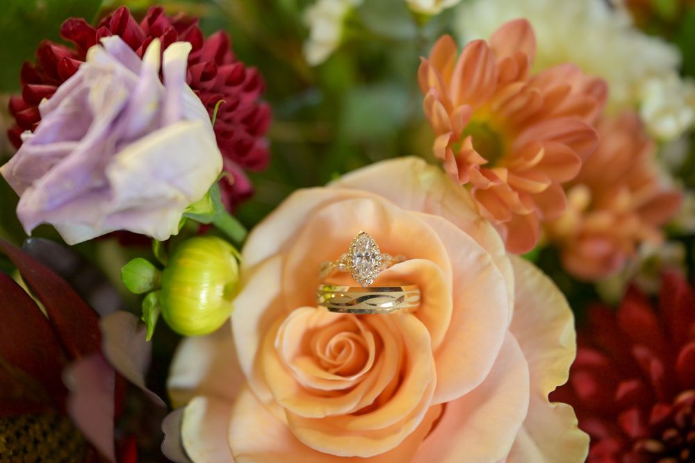 Diamond engagement ring and gold wedding band resting on a peach rose, surrounded by pink, coral and burgundy wedding flowers.