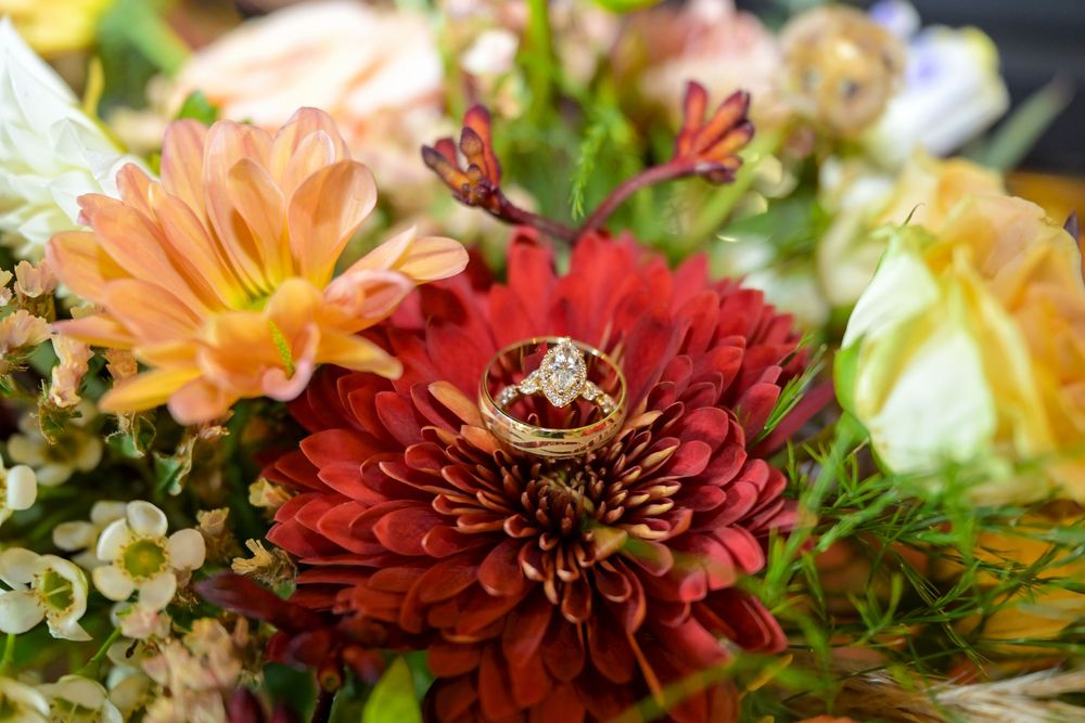 Wedding rings with a diamond engagement ring resting on a red dahlia surrounded by orange, peach and yellow bridal bouquet flowers.