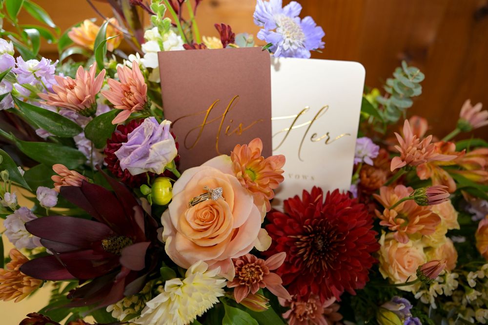 Wedding bouquet of peach roses, burgundy dahlias, and purple blooms with 'His' and 'Her' vow books tucked among the flowers.