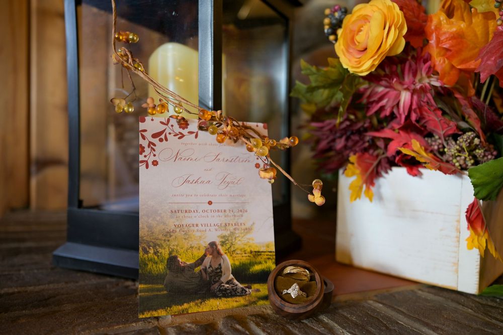 Autumn wedding invitation with couple's photo beside a lantern, rustic wooden box of orange and burgundy fall flowers, and a ring box.