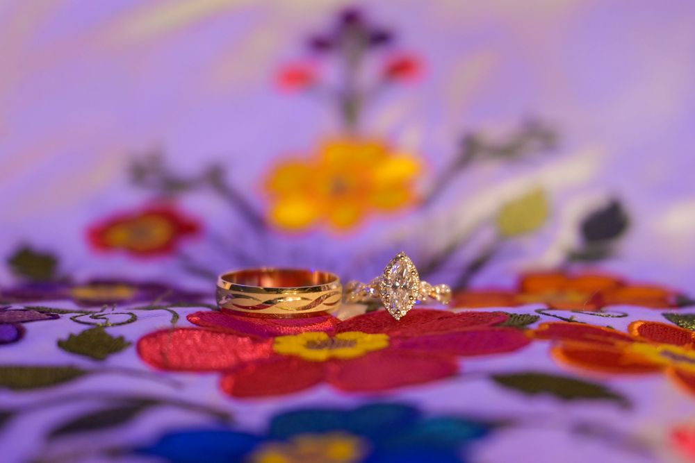 Gold wedding band and diamond engagement ring resting on colorful embroidered floral fabric with red, yellow, and green flowers.
