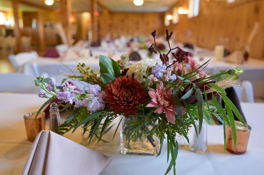 Floral centerpiece of burgundy dahlias, pink blooms and green foliage in a glass vase on a white-clothed table in a rustic wedding venue.