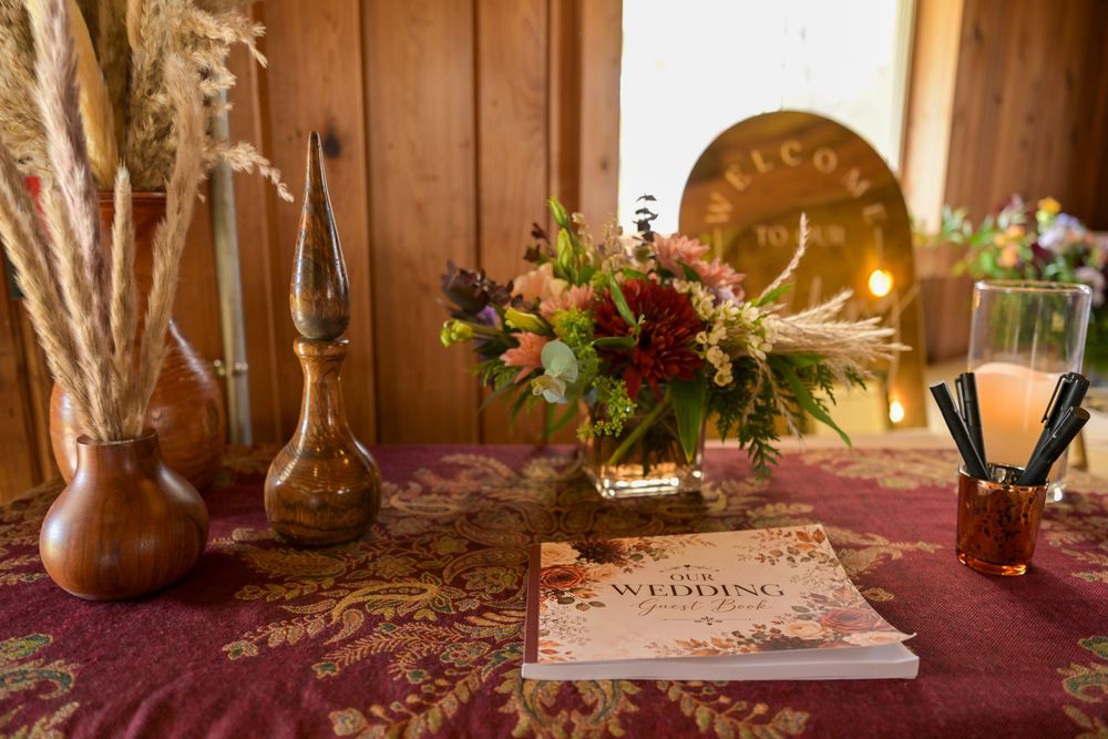 Rustic wedding guest book on a burgundy floral tablecloth beside an autumn flower arrangement, dried pampas grass, and brown glass bottles.