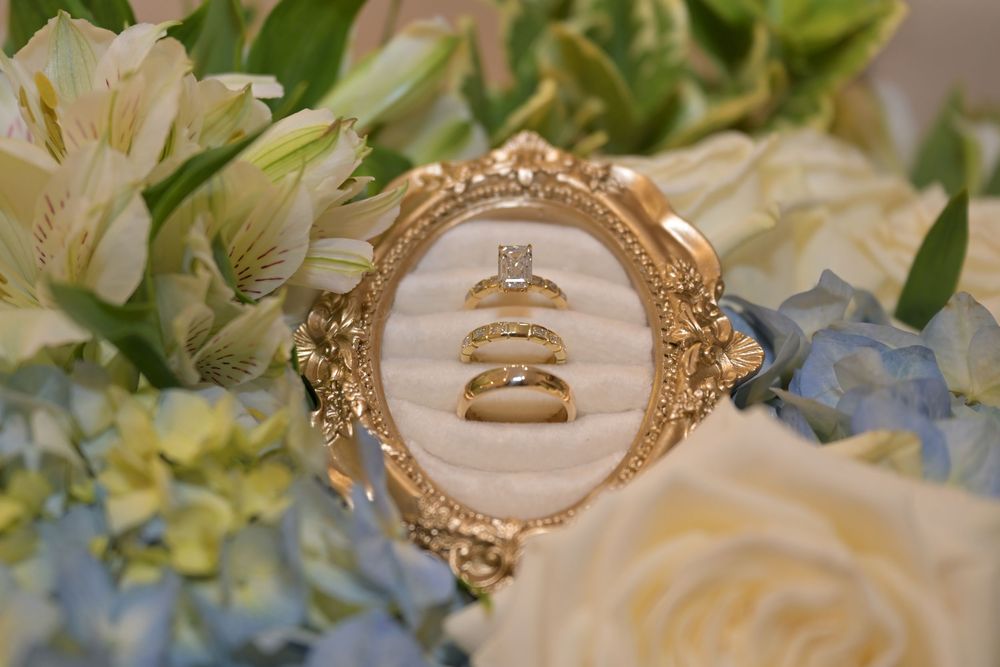 Gold ornate frame holding a diamond engagement ring and two wedding bands, surrounded by white roses and blue hydrangeas.