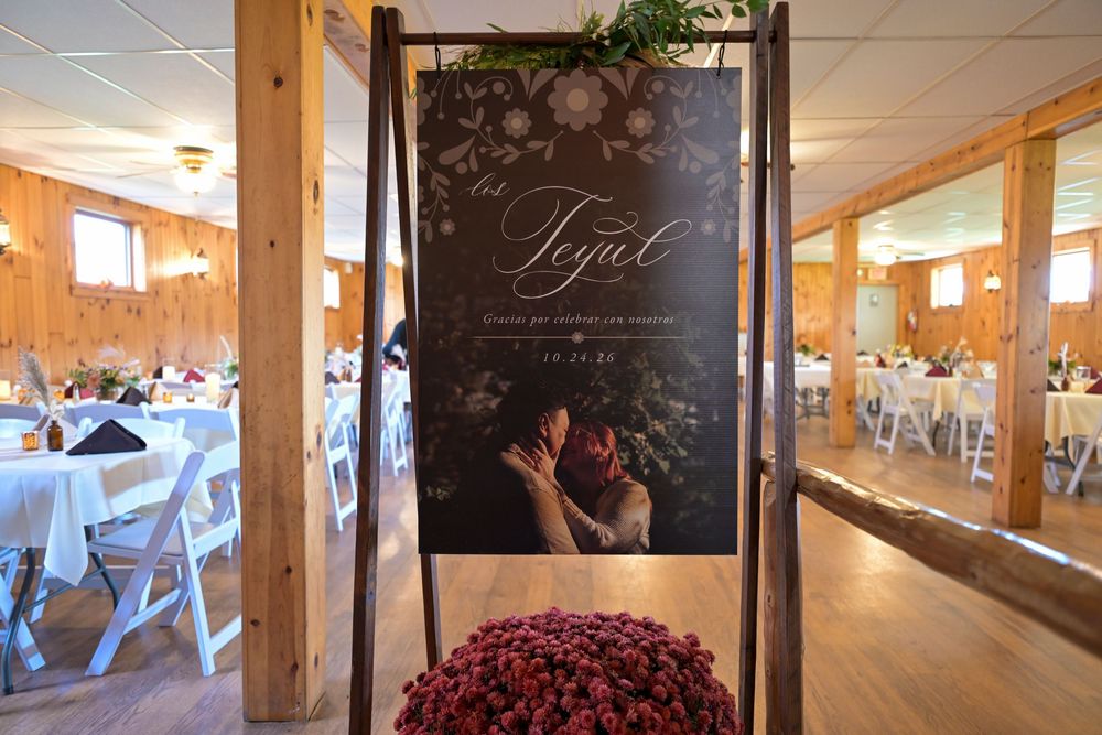 Rustic barn wedding venue with a floral welcome sign of a kissing couple, white tables and chairs, and a burgundy flower arrangement.