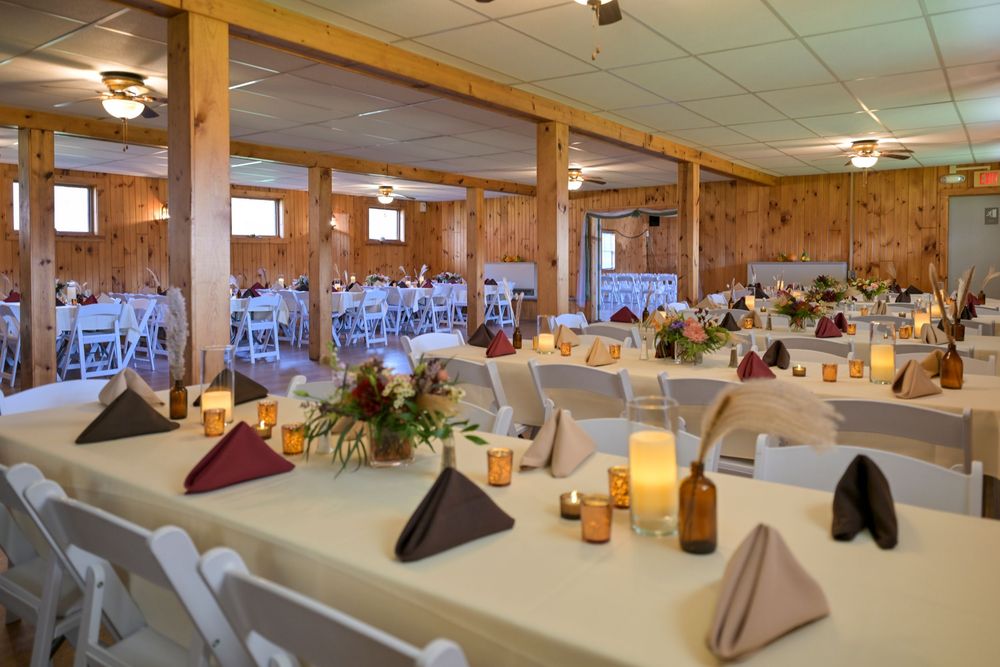 Rustic wooden banquet hall set for a wedding reception with long white tables, folded napkins, candles, floral centerpieces and chairs.