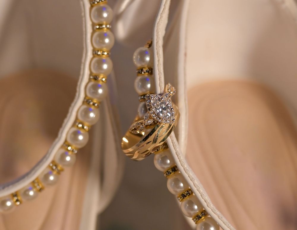 Gold diamond ring resting between ivory bridal shoes adorned with strands of pearl trim and gold accents.