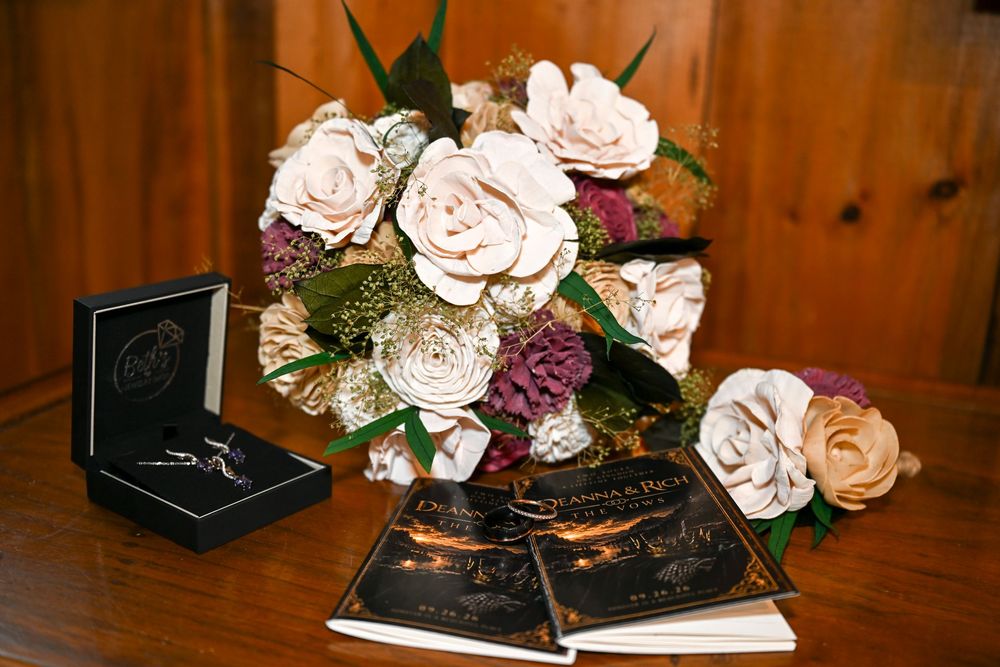 Rustic wooden flower bridal bouquet in blush, cream and plum beside an open jewelry box and wedding vow books on a wood table.