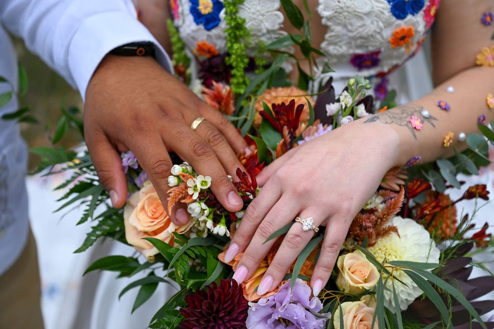 Bride and groom hold hands over a colorful bouquet of roses, dahlias and greenery, with wedding rings visible on their fingers.