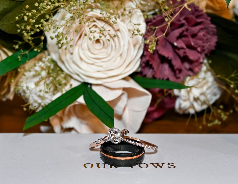 Diamond engagement ring and black and rose gold wedding band rest on a vow book in front of a cream and burgundy wedding bouquet.