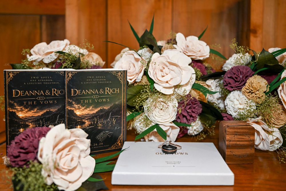 Two personalized wedding vow books titled Deanna & Rich beside a white keepsake box, surrounded by cream and mauve roses on a wooden table.