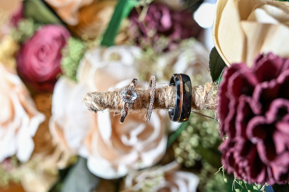 Wedding rings resting on twine-wrapped wood, surrounded by blush pink, cream and burgundy flowers in a rustic bridal bouquet.