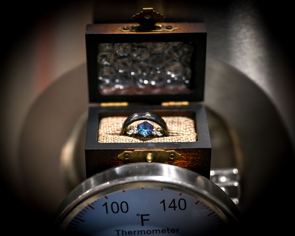 Unique black wedding band with blue gemstone displayed in wooden box atop vintage thermometer.