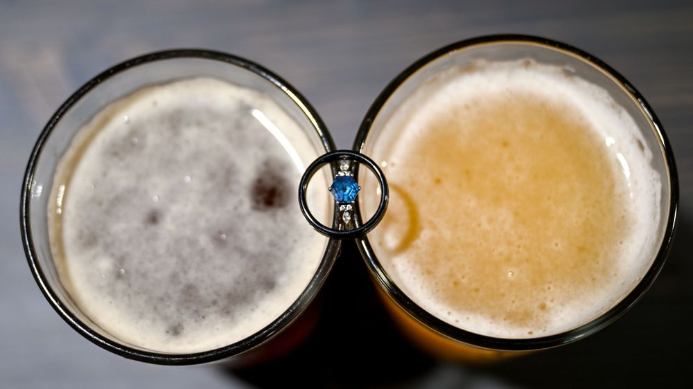 Two frothy beer glasses with a blue gemstone engagement ring balanced between them.