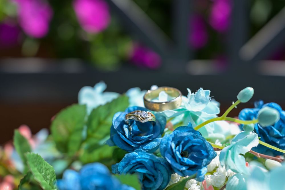 Gold wedding rings resting on vibrant blue roses in a colorful floral bouquet with green leaves and pink flowers in background.