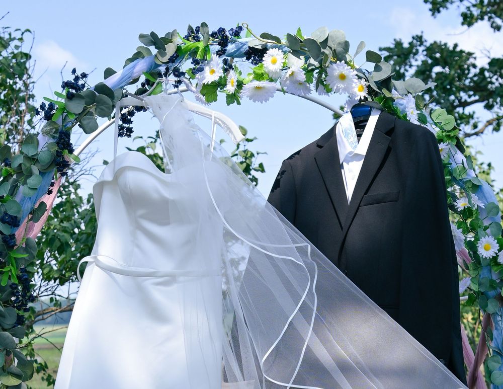 Wedding arch with floral decor displaying a white bridal gown with veil and black tuxedo outdoors.