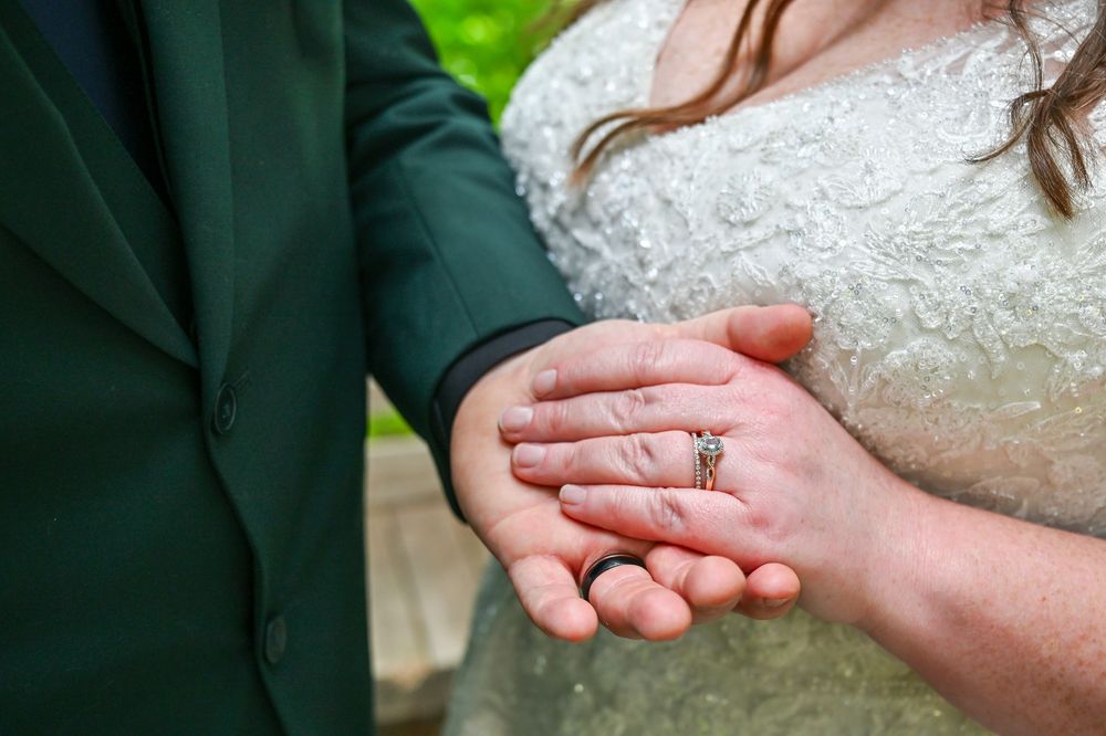 Close-up of a bride and groom's hands together showing a diamond engagement ring and wedding band, with a lace gown and green suit.
