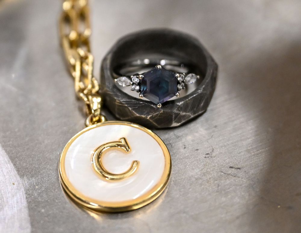 Gold initial C charm necklace beside a dark hammered ring with blue sapphire on silver surface.