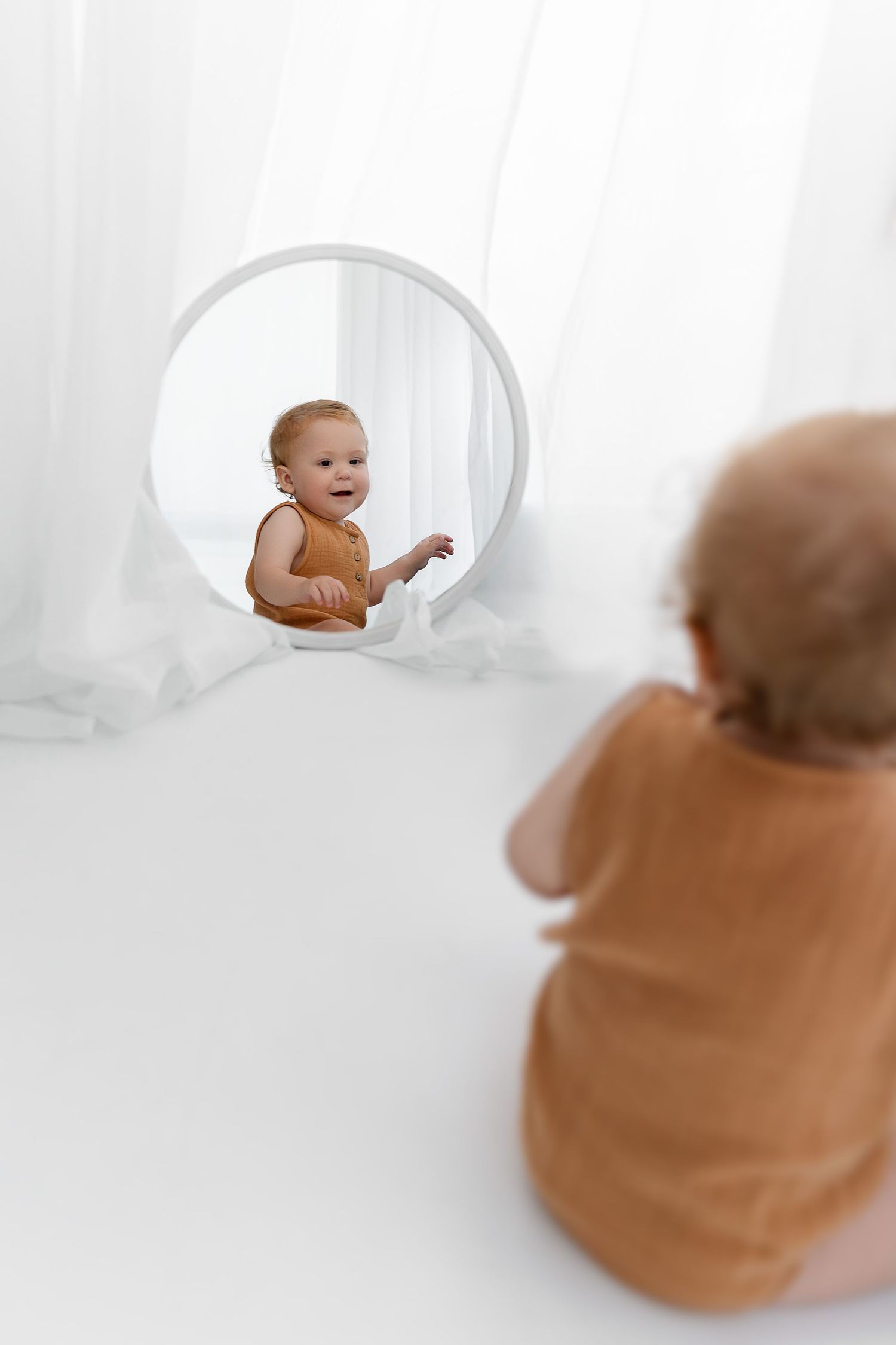 Baby sitting and looking at reflection in round mirror against white backdrop.