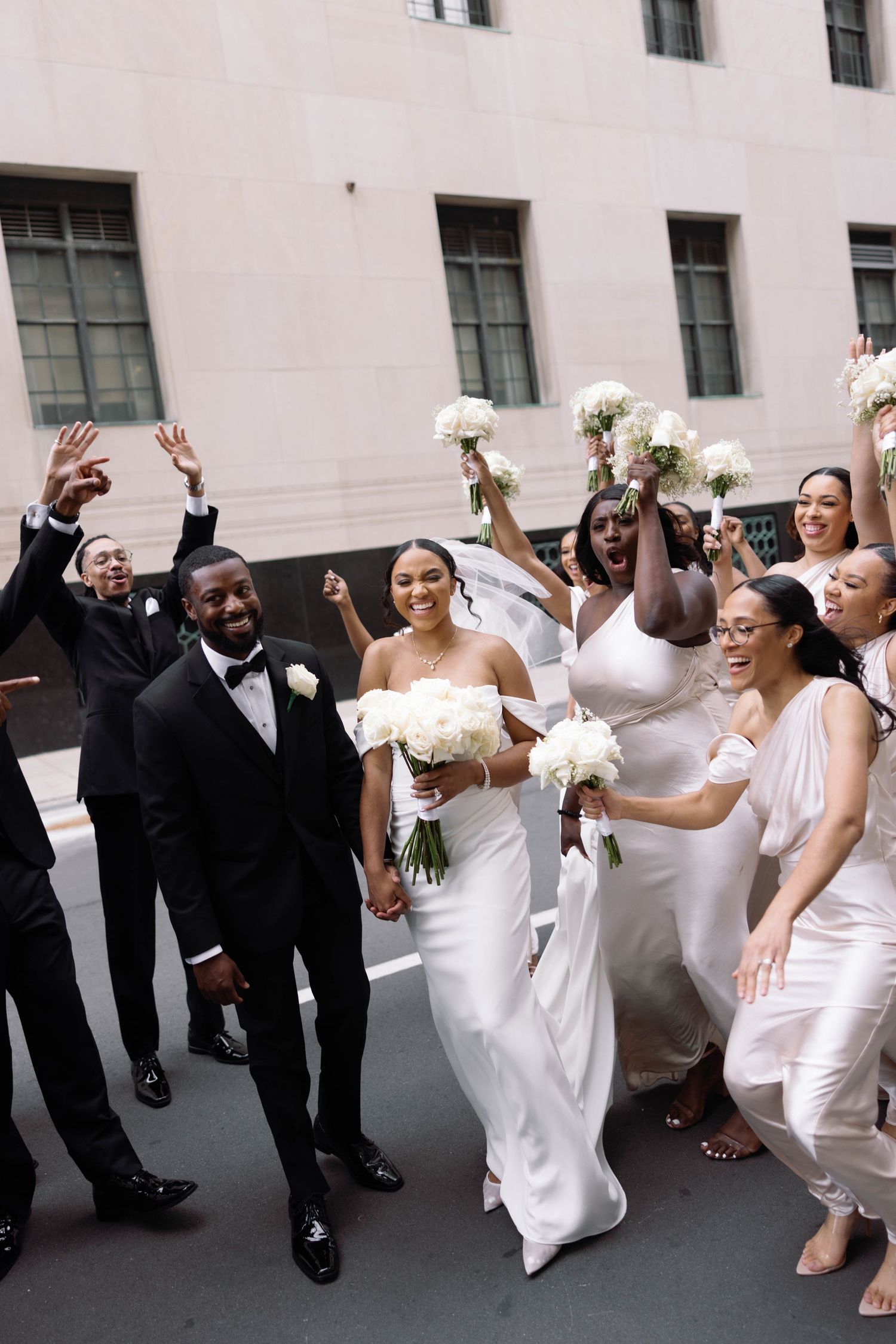 Wedding party celebrates joyfully on city street with bridesmaids in white dresses.