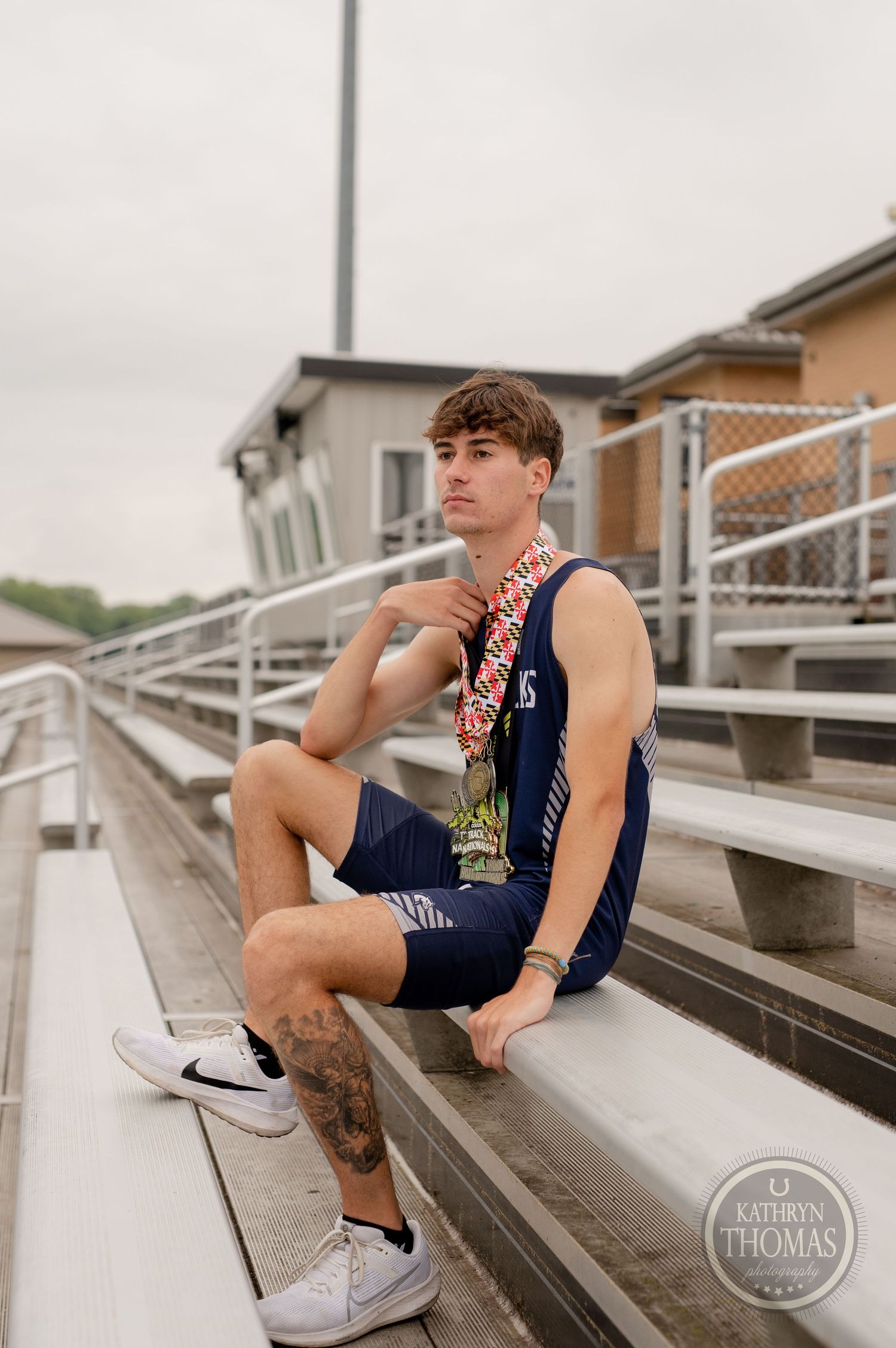 Student athlete in varsity letterman jacket sits on metal bleachers wearing athletic attire and sneakers.