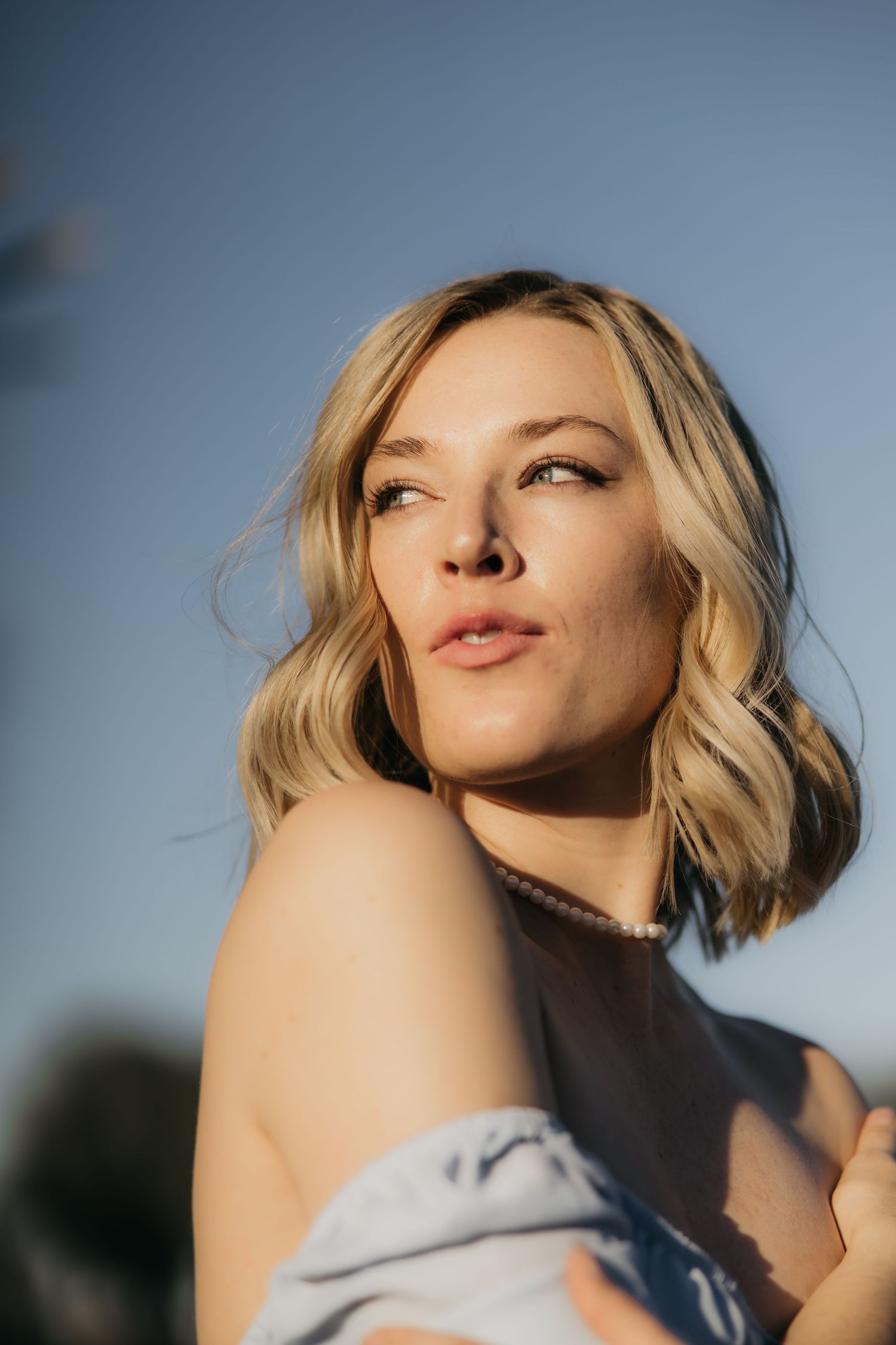 Close up golden hour portrait of a blonde woman with wavy hair looking upward against a blue sky background.
