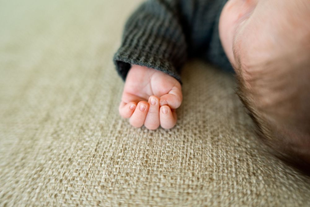 A tiny infant hand reaches out from a dark gray sweater sleeve against a beige textured fabric background.