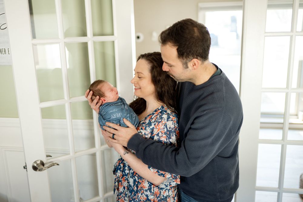 Adults share a tender moment with a newborn baby next to white French doors in a bright room.
