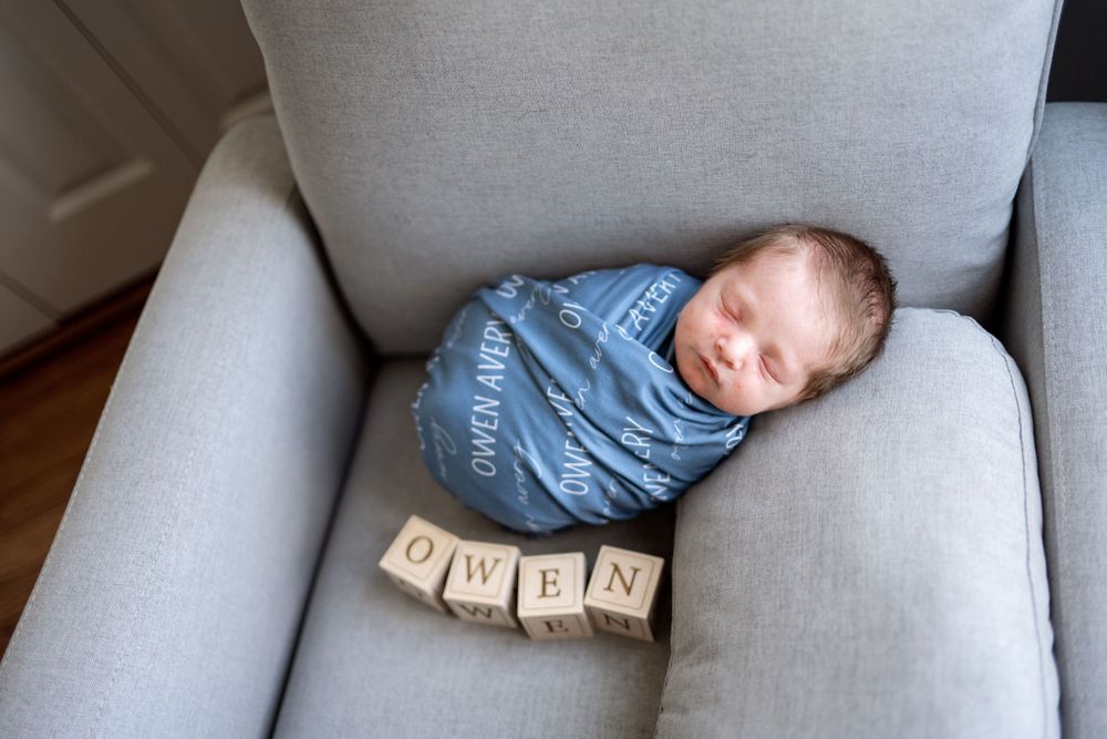 Sleeping baby swaddled in blue fabric lies on gray couch with wooden alphabet blocks spelling 'OWEN'.